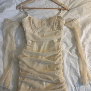 Champagnefärgad omlottklänning med mesh - Säljer en champagnefärgad kortklänning med omlottdetaljer och transparenta meshärmar. Klänningen har smala axelband, offshoulder-ärmar och rynkade partier längs hela klänningen för en snygg siluett. Jag har haft den på mig på min bal bara en gång, annars är den ny💓Perfekt för dig som gillar trendiga och romantiska plagg.