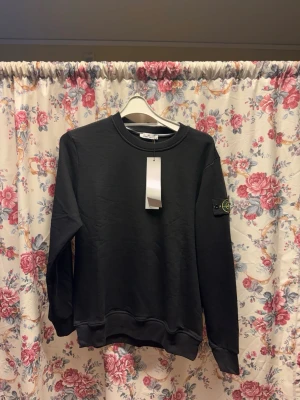 Svart sweatshirt från Stone Island - Har S-M-L       Svart långärmad sweatshirt från Stone Island i 100% bomull. Klassisk rund hals och ribbade muddar. Ikonisk Stone Island patch med broderad logga på vänster ärm. Perfekt för dig som gillar stilrena och exklusiva streetwear-plagg.