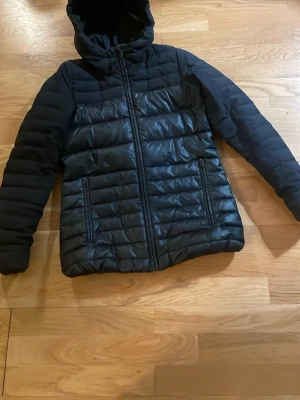 Svart pufferjacka med huva - Säljer en svart pufferjacka med huva och dragkedja framtill. Jackan har quiltad design på kroppen och släta ärmar, samt två fickor med dragkedja. Perfekt för kyliga dagar och enkel att matcha med olika outfits.