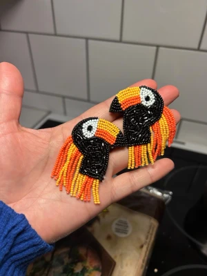 Färgglada papegojörhängen med pärlor - örhängen formade som papegojor, gjorda av små pärlor i svart, orange, gul och vit. Hängande pärlfransar ger extra färg och rörelse. Fäste med plupp på baksidan. Perfekt för dig som vill sticka ut med coola accessoarer. Material är tyg + pärlor och metall 
