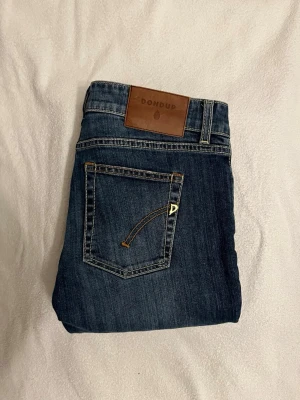 Dondup jeans  - Snygga blå jeans från Dondup i storlek 28. Klassisk femficksmodell med bruna detaljer och logopatch bak. Jeansen har raka ben och kontrastsömmar, tillverkade i en mix av bomull och polyester för skön passform. Perfekt för dig som gillar stilrena jeans med coola detaljer.