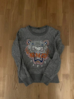 Grå Kenzo sweatshirt med tiger - Säljer en grå sweatshirt från Kenzo med det ikoniska broderade tigermotivet i orange, blått och vitt på bröstet. Tröjan har rund hals, långa ärmar och ribbade muddar. Perfekt för dig som gillar streetwear och vill sticka ut med ett coolt statement-plagg.