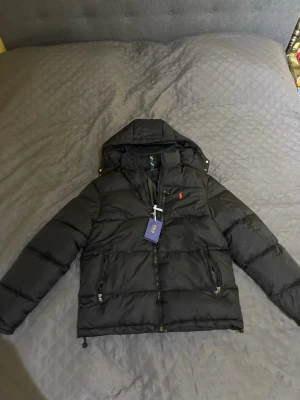 Svart pufferjacka från Polo Ralph Lauren - Svart pufferjacka från Polo Ralph Lauren med huva och dragkedja framtill. Jackan har två sidofickor med dragkedja och ett litet rött Polo-logo broderat på bröstet. Perfekt för kalla dagar och har en schysst, modern look.