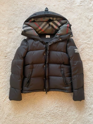 Svart pufferjacka från Burberry - Säljer en svart pufferjacka från Burberry med ikonisk rutig huva i grått, rött och vitt. Jackan har dragkedja, knappar och två fickor med dragkedja framtill. Märke med Burberry London England på ärmen. Perfekt för kalla dagar och riktigt snygg stil. Den är nästan helt ny använd några gånger 