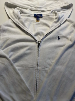 Ralph Lauren zip hoodie  - Säljer en vit zip hoodie från Ralph Lauren. Hoodien har dragkedja, fickor, huva och en marinblå logotyp. En mycket snygg hoodie som går att matcha med allt och passar för alla årstider. Säljer på grund av att den inte passar mig. Priset är inte hugget i sten!