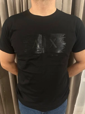 Svart Armani Exchange t-shirt - Snygg svart t-shirt från Armani Exchange med ett stilrent tryck i svart på bröstet där det står 'A|X'. T-shirten har rund hals och korta ärmar, perfekt för en clean och modern look. Materialet känns mjukt och bekvämt mot huden.
