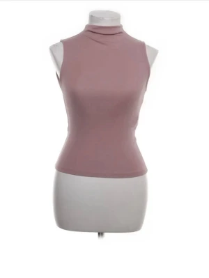 Ljusrosa polotopp ärmlös från Zara - Säljer en ljusrosa polotopp med ärmlös design och tight passform. Toppen har en hög krage och är tillverkad i mjukt syntetmaterial som sitter skönt mot kroppen. Perfekt att styla med jeans eller kjol för en clean look.