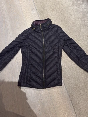 Svart dunjacka från Michael Kors - Snygg svart dunjacka från Michael Kors med quiltad design och guldfärgade dragkedjor. Jackan har hög krage och är lätt att packa med sig. Perfekt för dig som vill ha en stilren look under kyliga dagar.