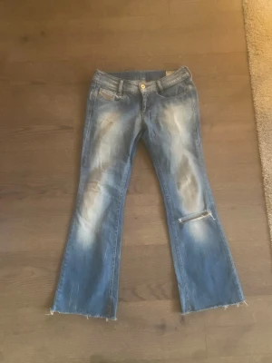Blå bootcut jeans från Diesel Louvely - Säljer ett par blå bootcut jeans från Diesel  med slitningar och råa kanter. Jeansen har en ljus tvätt, klassiska fem fickor och ett snyggt slitet hål på ena benet. Materialet är stretchigt jeans och de har en låg midja.