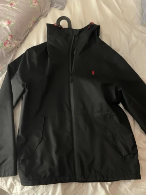 Svart vindjacka från Polo Ralph Lauren - Svart vindjacka från Polo Ralph Lauren med röd logga på bröstet och röd insida. Storleken är xs men sitter mer som S/M personen på bilden är 179 cm lång. Nypris är ca 2300 kr. Jackan är i grymt bra skick och omhändertagen väl. Hör av er vid frågor!