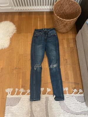 Blå jeans från 157 med slitningar - Säljer ett par blå jeans från 157 med skinny passform och snygga slitningar vid knäna. Klassisk femficksmodell med brunt läderpatch bak i midjan. Jeansen har hög midja och är gjorda i jeansmaterial med gul kontrastsöm. Perfekt för dig som gillar en cool och avslappnad stil.