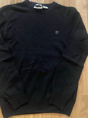 Stickad tröja från Timberland - Svart stickad tröja från Timberland med rund halsringning och diskret logga broderad på bröstet. Mjuk känsla och klassisk passform, perfekt för kyliga dagar då den är mjuk och skön