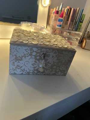 Beige smyckesbox med pärldetaljer - Säljer en snygg smyckesbox i beige med vitt spetsmönster och pärlemordetaljer på locket. Insidan har fack för ringar och smycken, och locket pryds av dekorativa pärlor och snören. Perfekt för att hålla koll på dina favoritsmycken.