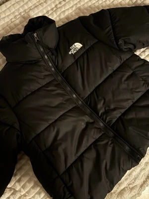 Svart pufferjacka från The North Face - Säljer en svart pufferjacka från The North Face med hög krage och dragkedja framtill. Det är den kortare varianten. Använd men i väldigt bra skick Passar M/L.