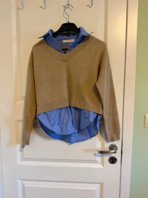 Beige stickad tröja med blå skjorta - Snygg beige stickad tröja från Mango med v-ringad hals och långärmad passform. Undertröjan är en blå skjorta som ger en cool lager-på-lager look. Perfekt för dig som gillar att mixa klassiskt med trendigt. Tröjan är loose fit. Detaljerna från skjortan är fastsydda.