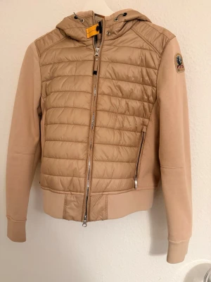 Beige pufferjacka från Parajumpers - Snygg beige/ champagne färgad pufferjacka från Parajumpers med huva och coolt gult band vid halsen. Jackan har quiltad framsida, dragkedja och fleeceärmar med patch på axeln. Perfekt för dig som vill ha en stilren och bekväm jacka med street vibe. Nästintill aldrig använd.