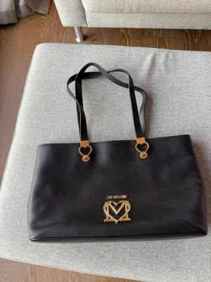 Svart handväska från Love Moschino - Snygg svart handväska från Love Moschino med guldfärgade hjärtformade detaljer på axelremmarna och ett stort guldfärgat logomärke framtill. Insidan är fodrad i starkt röd färg med innerfack och dragkedja. Väskan är tillverkad i skinn och har en stilren, rektangulär form.