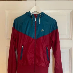 Nike vindjacka röd och blå S - Nike vindjacka i rött och blått med huva och vita snören. Jackan har dragkedja framtill, två fickor med dragkedja och klassisk Nike-logga på bröstet. Materialet är lätt polyester och jackan har en sportig look med kontrastfärger. Litet slitage märke på framsidan, se bild. Annars i bra skick.