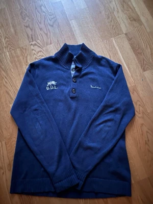 Marinblå Bondelid polotröja L - Snygg marinblå polotröja från Bondelid med broderad logga och B.D.L. på bröstet. Tröjan har ribbad krage, tre knappar framtill och långa ärmar. Materialet är mjukt och stickat, perfekt för kyliga dagar. Passar dig som gillar klassisk och stilren look. Vintage look, liknande till hollister. Storlek L hör av dig gällande frågor 