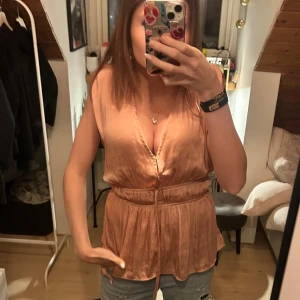 Persikofärgad / rosa lager omlottblus med knyt - Säljer en persikofärgad omlottblus i mjukt, glansigt material. Blusen är ärmlös med djup v-ringning och har ett smalt knytband i midjan som ger en snygg siluett. Passformen är loose och toppen har ett lätt plisserat fall.