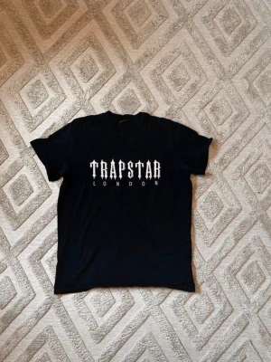 Svart Trapstar London t-shirt - Säljer en stilren svart t-shirt från Trapstar London. T-shirten är i utmärkt skick och perfekt för en snygg look. Den är i storlek M men kan också passa storlek S. Jag är aktiv 24/7 och svarar gärna på alla frågor. Frakten kan göras samma dag eller nästa dag, så du får din t-shirt snabbt! Missa inte chansen att köpa! Tveka inte att höra av dig!
