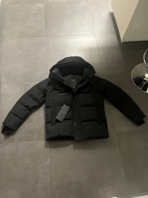 Canada Goose Wyndham black badge - Säljer en svart dunjacka från Canada Goose i storlek M. Jackan har huva, diskret logga på ärmen och stängs med både dragkedja och knappar. Fodrad för extra värme och perfekt för kalla dagar. Klassisk, stilren design som funkar till allt.