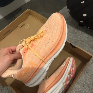Hoka sneakers i ljus orange - Säljer ett par  Hoka sneakers i ljus orange med vita och persikofärgade detaljer. De är HELT nya och kartongen finns även.