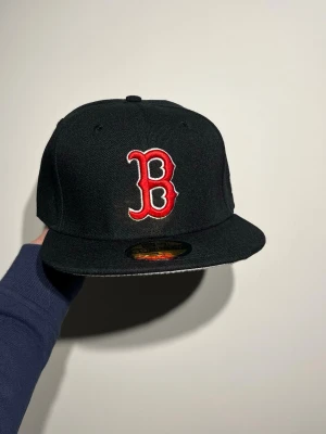 Svart Boston Red New Era keps - Säljer en svart New Era 59FIFTY keps med Boston Red Sox logga broderad i rött och vitt framtill. Kepsen har en platt skärm i grått och MLB-logga baktill. Passar dig som gillar sportig streetstyle och klassiska baseballkepsar.