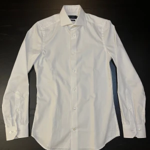 Vit super slim fit skjorta från Riley - Säljer en stilren vit skjorta från Riley i super slim fit-modell. Skjortan har klassisk krage, långa ärmar och knappar framtill. Perfekt för dig som gillar en tajt och modern passform. Materialet är mjukt och känns skönt mot huden.