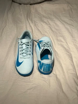 Nike Mercurial fotbollsskor blå/vit - Säljer ett par Nike Mercurial fotbollsskor i storlek 40. Skorna är vita med blå detaljer och har en platt sula som är perfekt för spel på inomhusplan. Snygg design med stort Nike-logga på sidan och Mercurial-tryck på hälen. Tillverkade i syntetmaterial med snörning framtill.