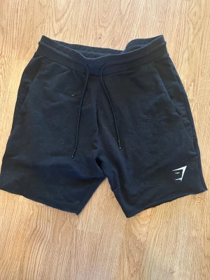 Svarta shorts från Gymshark med snörning - Svarta shorts från Gymshark med stilren logga på vänster ben. Mjukt bomullsmaterial och bekväm midwaist-passform. Utrustade med snörning i midjan för extra justering. Perfekta för träning eller chill dagar.