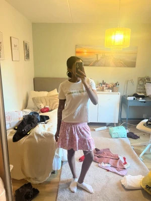 Rosa blommig kortkjol med volang - Supersöt kortkjol med blommigt mönster i rosa och vitt. Kjolen har en smockad midja och volang nertill som ger en flowy känsla. Perfekt för varma dagar och lätt att matcha med en enkel t-shirt eller linne. Storlek S från Ginatricot, aldrig använd men utan prislapp.