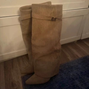 Beiga boots med klack - Snygga höga boots i beige mocka med spetsig tå och guldfärgad spänndetalj upptill, super fina men får inte till användning därav jag säljer. På en av skorna har ena grejen som spänner fast snörena gått av, däremot inget som störs när man har på sig dom!
