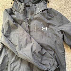 Grå vindjacka från Under Armour - Snygg grå vindjacka från Under Armour med huva och dragkedja. Jackan har logga på bröstet, meshfoder och praktiska fickor med dragkedja. Perfekt för blåsiga dagar och outdoor-aktiviteter. Sportig look och bekväm passform.