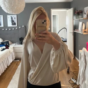 Beige omlottopp från Gina Tricot - Säljer en beige omlottopp från Gina Tricot med långa puffiga ärmar, djup v-ringning och en lös passform