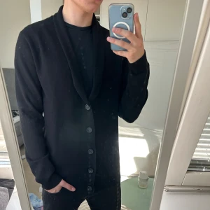 Lindbergh Svart kofta  - Svart kofta från Lindbergh i relaxed fit med snygg sjalkrage och knäppning framtill. Långärmad och enkel design som passar perfekt till lager-på-lager. Mjuk och skön, perfekt för kyligare dagar.