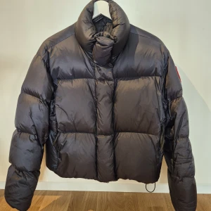 Canada Goose jacka helt ny - Säljer en svart, riktigt fet dunjacka från Canada Goose med klassisk logga på ärmen. Jackan är puffer-modell med hög krage och breda kanaler, perfekt för kalla dagar. Materialet är glansigt och jackan har en loose fit som ger en cool siluett. Jackan är helt ny!