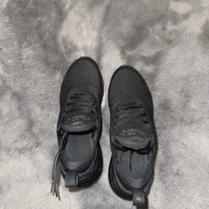 Svarta Nike Air Max 270 sneakers - Säljer ett par helsvarta Nike Air Max 270 sneakers med snygg mesh-ovandel och tydlig Air Max-dämpning i hälen. Skorna har snörning och en sportig vibe, perfekta för dig som gillar streetstyle och komfort. Diskret Nike-logga på sidan och plös.