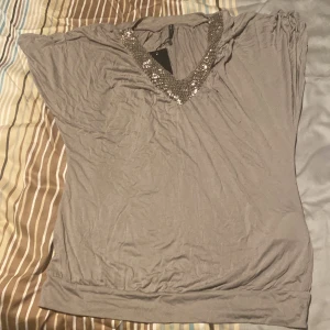 Grå/beige  topp med paljettdetaljer från Amisu - Snygg grå topp från Amisu i lös passform med korta ärmar. V-ringad hals med bred kant täckt av silvriga paljetter som ger extra bling. Mjuk och skön i bomullsmix, perfekt när du vill sticka ut lite extra. Enkel att matcha med både jeans och kjol.