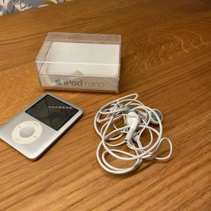 iPod nano - Apple iPod nano i silver med originalförpackning och hörlurar. Enheten har klassisk klickhjul och kompakt design. Skärmen och höljet ser ut att vara i gott skick, inga tydliga repor eller skador syns. Perfekt för musikälskare som vill ha en smidig MP3-spelare.