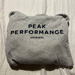 Grå hoodie från Peak Performance - Snygg grå hoodie från Peak Performance med svart broderad logga på bröstet. Klassisk känguruficka framtill, ribbade muddar och skön huva. Perfekt för chill dagar eller när du vill ha en enkel och stilren look. I storlek 160cm