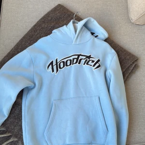 Ljusblå hoodie från Hoodrich - Säljer en ljusblå hoodie från Hoodrich med stor svartvit logga framtill och broderad detalj på huvan. Tröjan har känguruficka, huva och långa ärmar. Perfekt för dig som gillar streetwear och vill sticka ut med en snygg färg.
