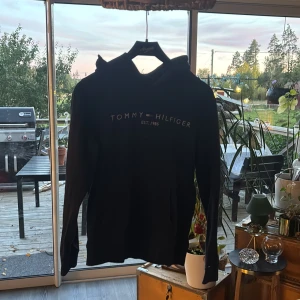 Svart hoodie från Tommy Hilfiger - Marinblå  hoodie från Tommy Hilfiger med broderad logga och text 'EST. 1985' på bröstet. Klassisk huva och långärmad modell i mjuk bomullsmix. Perfekt för dig som gillar stilrena och bekväma plagg med ikonisk branding.