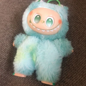 Söt fluffig labubuhandväska - Unik handväska formad som ett gulligt labubu med stort leende och gröna ögon. Väskan är täckt av mjuk, ljusblå och mintgrön päls med små öron och handtag upptill. Perfekt för dig som vill sticka ut med en lekfull accessoar!