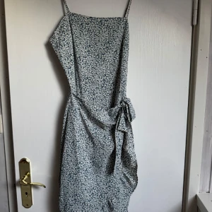 Blommig omlottklänning med knyt - Söt långklänning med smala axelband och blommigt mönster i vitt och blått. Klänningen har omlottmodell med knyt i midjan och en lätt, flowy känsla. Perfekt för varma dagar när du vill känna dig både chill och snygg.