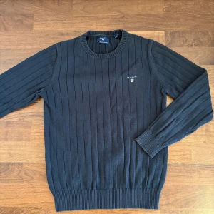 Svart ribbstickad tröja från Gant - Svart ribbstickad tröja från Gant i bomull med rund halsringning och diskret logga på bröstet. Klassisk modell med långa ärmar och breda muddar vid ärmslut och nederkant. 