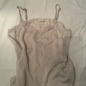 Beige linnetopp med spets från JDY - Supersöt beige linnetopp från JDY med tunna axelband och v-ringning dekorerad med spets. Toppens material känns lätt och flowy, perfekt för varma dagar eller lager på lager. Enkel och stilren design som är lätt att matcha.