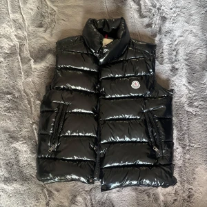 Monclear Tib Gilet väst  - Monclear Tib Gilet dunväst från Moncler med glansig finish och klassisk logga på bröstet. Västen har hög krage, två sidofickor med dragkedja och quiltad design. Stängs med både dragkedja och tryckknappar. Storlek 1 (motsvara S) inköpt 2021. Mycket bra skick. 
