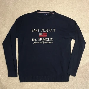 Mörkblå stickad tröja från Gant - Mörkblå stickad tröja från Gant med broderad amerikansk flagga och text på bröstet. Passar perfekt nu till hösten! Vid minsta fråga eller fundering är det bara att höra av er!🤝🏼