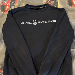 Mörkblå Sail Racing tröja - Mörkblå tröja från Sail Racing med broderad vit logga på bröstet. Klassisk rund halsringning och långa ärmar. Tillverkad i mjuk bomull, perfekt för en avslappnad stil.
